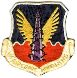 a shield, top half is blue, bottom half red, on it is a grey mololith wrapped in barbed wire with gold eagle wings spread from behind, there is a banner across the bottom with first air commando wing written on it