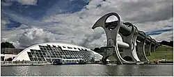 The Falkirk Wheel dominates the right of the picture with the tourist shop and restaurant dwarfed by the Wheel on the left, the bottom shows the marina with a boat in the centre
