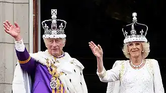 Charles and Camilla wearing their crowns and coronation robes waving from the balcony of Buckingham Palace