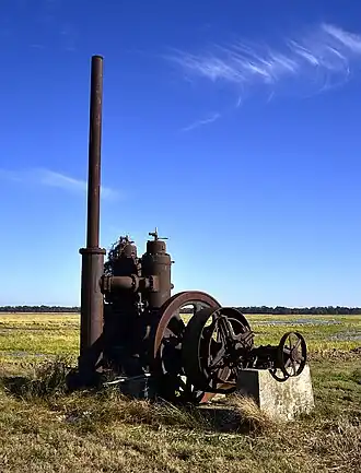 Photo of a very rusted industrial-looking pump with tall tube and wheel sits in a flat rice field.