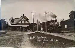 A postcard of a passenger train with a steam locomotive at a two-story wooden station