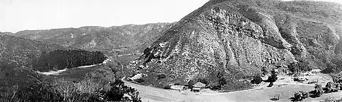 Black and white panoramic view of a canyon, a group of trees is to the left and several residential buildings are at lower right.