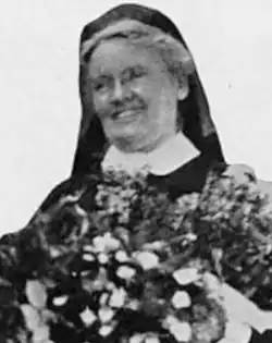 A smiling older white woman wearing a head covering, eyeglasses, and a high starched collar; there is a bouquet in the photo's foreground