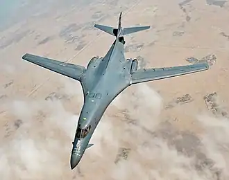 Top view of B-1B in-flight with white clouds scattered underneath. Its wings are swept 20 degrees short of fully forward.