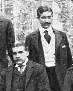 Sullivan, seated, with Herbert standing behind his left shoulder; both are very well-dressed and mustachioed