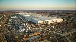 Photo is of a large scale manufacturing facility under construction, captured from above with aerial photography. There are many cranes, heavy equipment, materials, and other machinery surrounding the tall plain walls of the main facility.