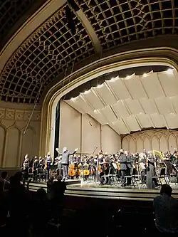 In a ivory concert hall, a stage full of classical musicians and conductor are standing at the end of a performance