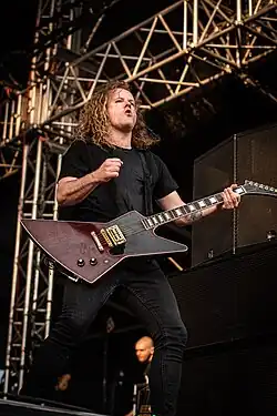 Tyrrell plays guitar with both hands. He has a black T-shirt and black jeans with a tattoo on his right hand.