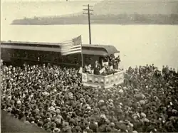 A dramatic political scene. Beside a river stands a podium, on which a flagpole flies a huge American flag. Beneath the flag stands a candidate in a dark suit addressing an impressive crowd which takes up most of the photograph. Not only the quayside but a ferry beside it on the water are packed full of people listening intently.