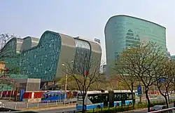 Two irregularly shaped greenish glass-faced buildings rise over a street lined with freshly budding trees on which a blue and white trolleybus is stopped.