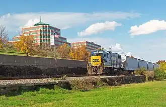 A blue diesel locomotive, with "CSX" in blue letters on its yellow front, pulling several hopper cars along a railroad track. In the background are some postmodernist brick office buildings with green spired roofs.