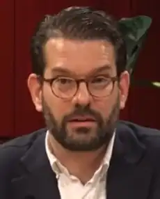 A caucasian man with a beard and glasses wearing a suit indoors