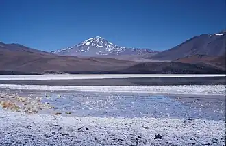 Mountains, including a snow-covered one in the middle, rise above a white surface with a lake