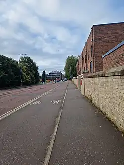 A street with a brick building to the right side, separated from the street by a stone wall. There are additional buildings and cars further down the street.