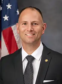 Official portrait of Chase Tramont. He is a mostly bald, middle-aged, white man wearing a suit and blue tie. Behind him is the French flag partially visible.