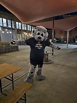 A mascot in the shape of an anthropomorphic harbor seal standing with its hands to the sides, and wearing a dark blue shirt saying "Never say die"