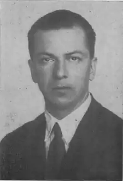 A young man in a dark suit and tie, with a serious expression.