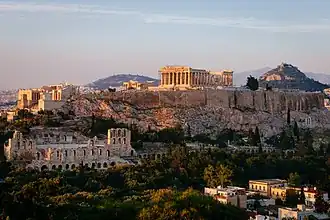 Color photograph of a cityscape, with a slightly forested landscape in the foreground, a rock face in the middle and rolling hills in the background. Ruins of large Classical buildings can be seen in the foreground and middle,