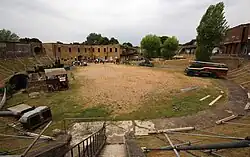 Wide view of the fort's interior, showing a semi-circular sandy area in which items of military equipment are standing. A row of brick buildings is visible at the rear.