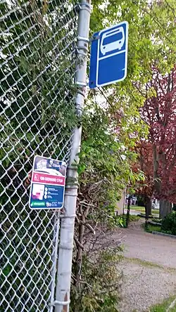 a bus stop sign for Cobourg Transit, attached to a pole holding up a fence of a elementary school playground. On the fence next to it is another sign advertising Cobourg Ride, an on demand transit pilot. There are plenty of differently coloured trees in the background of the image.