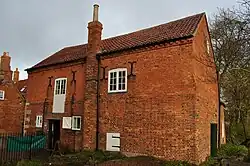A general view of the mill building from the bridge, taken with a wide angle lens, resulting in some false verticals. The shot is tricky, but it is probably the only way to capture the whole building without obscuring trees. it is November, and the low autumn sun makes the red brick appear to have an orange glow. We see a two-story building in 18th century red brick, with a pitched roof clad in pantiles of nearly the same colour. A narrow chimney breast breaks the flatness of the wall in the centre, and the chimney continues above the eaves and is capped with an unusually tall plain earthenware pot, in a pale yellow colour characteristic of the area. To the right of the chimney, in the wall above the millrace, the wall is pierced only by a small white painted door on the ground floor and a double window on the first. On the left of the chimney, however, there are several white-painted openings: a door and two windows on the ground floor, and a tall door on the second, for the grain hoist access. Four black painted iron scrolls terminate four threaded tie-bars through the building at what would appear to be head height on the second floor. To the left (north) of the mill is a second building, the mill cottages, and these can be made out on the extreme edge of the picture. They feature stone quoins in their red brick wall, and the cornder of the mill next to them has stone quoins on the ground floor too.