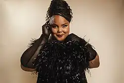 Pinnock smiles directly at the camera. She wears sheer black gloves and a feathered and sequined dress. Her hair is in box braids pulled back in an updo, and one hand pushes her hair behind her ear.