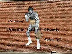 A red brick wall with a grey-scale mural of a boxer. He is wearing a golden championship belt on his waist and a golden plaque is by his left knee. Over the image are painted the words "The Home of Demetrius "Oaktree" Edwards, Ayden, NC".