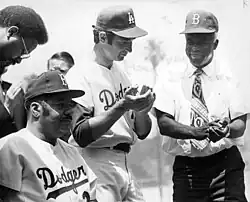 three men wearing baseball caps, two standing and clapping, one seated and looking ahead.