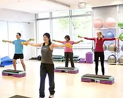 Four people exercise with dumbbells in the hands, standing behind plastic step tools.