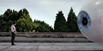 A man stands on a rooftop across from a giant eyeball.