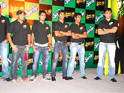 A group of men standing, wearing black T-shirts and blue faded jeans. All of them are looking to the left of the image. The backdrop has alternate black and green boxes.