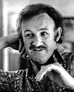 Black-and-white portrait of a middle-aged Hackman with dark hair and moustache, wearing a patterned collar shirt, resting his hand on his head and looking over to the side.