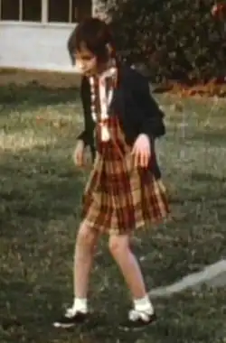 A slightly blurry color picture of Genie, facing slightly right of the camera, walking by herself outside in the hospital recreation yard. She is wearing a red and orange plaid-patterned dress and a thin blue sweater, with white socks and black shoes, and looks extremely pale, emaciated, and expressionless. Her limbs are exposed and look extremely thin. Both of her knees are very bent, and her arms are bent forward with both hands hanging down as she holds them out in front of her.