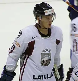 An ice hockey player standing, looking to the right of the camera. He is wearing a black helmet and a white uniform.