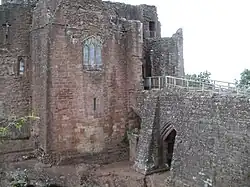 A castle, with a flat fronted tower facing the viewer with a stained glass window in the middle of it; a stone causeway is on the right of the picture, leading to a gateway to the right of the tower – a partially filled arch is supporting the causeway.
