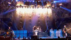 A band performing live. Two men play guitars, one to left and one to the right and one man in between the two guitarists sings into a microphone. One man is playing drums behind the other three men.