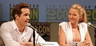 Reynolds speaking into a microphone sitting next Blake Lively, who is smiling