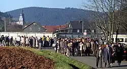 A large number of people of various ages standing and walking along a road in front of a high concrete wall, behind which houses and a church are visible in a wooded valley.