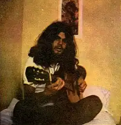 A man with long, black hair holding a guitar, sitting cross-legged