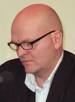 bald white male in profile, wearing glasses, white shirt and dark jacket, looking down, with a microphone in front