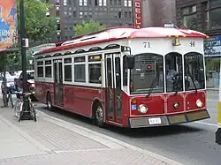 a bus adjacent to a large sidewalk in an urban area, it is red on the underside and white on the upper half, with a red roof. it seems slightly dated in design style, and has two doors on one side.