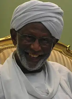This photograph features the late Hassan al-Turabi with a warm smile and a neatly groomed beard, wearing a white turban and traditional Sudanese attire. He is also wearing glasses, which frame his cheerful and kind expression. The background shows part of a golden, ornate chair with striped upholstery, adding a touch of elegance to the setting.