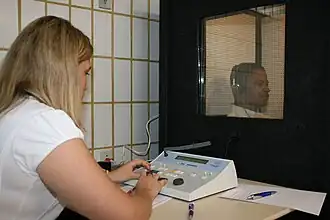 a female medical professional is seated in front of a special sound-proof booth with a glass window, controlling diagnostic test equipment. Inside the booth, a middle-aged man can be seen wearing headphones and is looking straight ahead of himself, not at the audiologist, and appears to be concentrating on hearing something