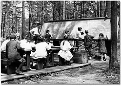 group of children in an outdoor classroom seated at desks and standing at blackboard