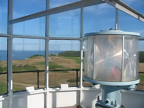 Photograph taken from the lantern room of the lighthouse. In the foreground on the right is the circular lens surrounding the lamp. The background is the view looking out over the lighthouse grounds and the cliff out to the ocean.