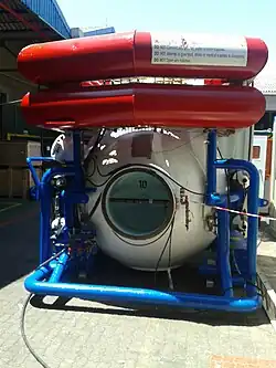 End view of a small hyperbaric chamber with open pressure door supported by a steel tubular frame, with two tubular ring floats at the top