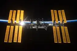 An image of the International Space Station. The silver-colored center module is dark blue, surrounded by four golden solar arrays on each side. The sun is reflecting off of the set to the left. In the background is the outline of the Earth.