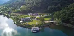 Aerial shot of a lakeside property showing one house built over a lake, a second house behind that with a large yard in front of it, and a third house behind the second, located on a slight incline of land, surrounded by mountains