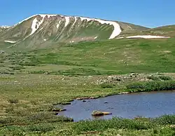 A grassy yet barren landscape with a small pond in the right foreground and distant ridgeline and mountains in the background