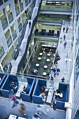 The image shows the inside of the Manchester Metropolitan University Business School. The image is taken from a corner, showing several floors within the building. You can clearly see staff and students walk around the building.